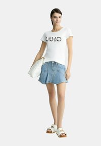 T-shirt bianca a maniche corte con logo "LIU.JO", abbinata a una gonna di jeans con orlo svasato. Gli accessori comprendono una borsa bianca e sandali.