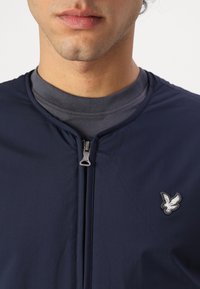 Veste bomber légère bleu marine avec fermeture éclair sur le devant, ornée d'un petit logo d'aigle brodé sur la poitrine gauche et d'une texture douce.