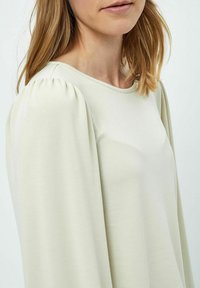 Maglietta beige chiaro a maniche lunghe con spalle a palloncino morbide e scollatura tondeggiante; realizzata in tessuto leggero e liscio.