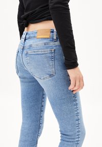 Jean en denim bleu clair avec une coupe slim, présentant des poches arrière et une étiquette en cuir tan au niveau de la taille. Le tissu a une texture délavée.