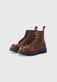 Botine din piele maro cu șireturi, cu tălpi negre și bucle de prindere, finisaj texturat, așezate pe un fundal gri deschis uniform.