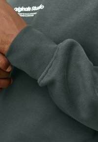 Una mano di carnagione scura che regola il polsino di un sudore verde scuro con del testo bianco parzialmente visibile sul petto.