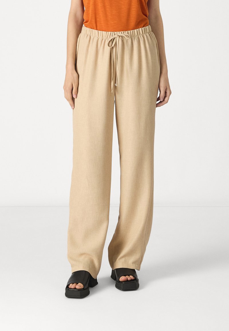 Pantalons larges beiges avec une taille à cordon, fabriqués dans un tissu texturé. Associés à des sandales noires à bouts ouverts.