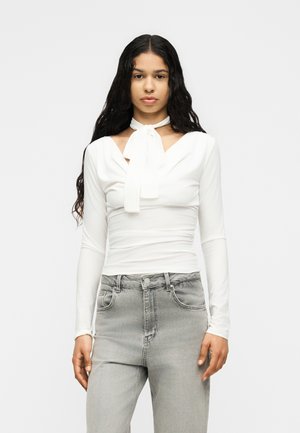 Woman with long curly hair wearing a white long-sleeve top with a neck tie and light gray high-waisted jeans, facing front.