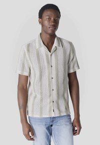 Jeune homme portant un pull à manches courtes à rayures verticales et un jean bleu clair, debout devant un fond clair uni.