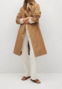 Manteau trench beige à double boutonnage, avec de grands boutons et des manches ceinturées, porté avec un pantalon crème et des sandales marron.