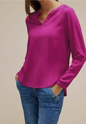 Fuksjowa bluzka z długim rękawem i dekoltem w kształcie litery V o miękkiej fakturze, w połączeniu z jasnoniebieskimi dżinsami z przednią kieszenią na zamek.
