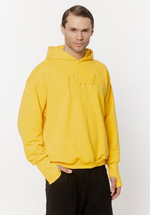 Mężczyzna w jaskrawożółtej bluzie z kapturem z nadrukiem, stojący z jedną ręką w czarnej kieszeni spodni na jednolitym białym tle.