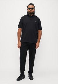 Čierna polo košeľa s krátkymi rukávmi, s jemným logom, v kombinácii s čiernymi nohavicami a zodpovedajúcimi teniskami, umiestnená na bielom pozadí.