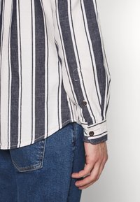 Camicia a righe bianca e blu navy, con maniche lunghe, orlo rifinito e dettagli in marrone sui bottoni dei polsini. Indossata con jeans blu.