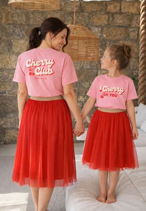 Frau und Mädchen halten Händchen, tragen passende rosa T-Shirts und rote Tüllröcke und lächeln sich drinnen an einer Steinmauer an.