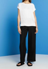 Femme portant un haut blanc uni à manches courtes, un pantalon noir à jambes larges et des sandales noires à bouts ouverts sur un fond bleu.