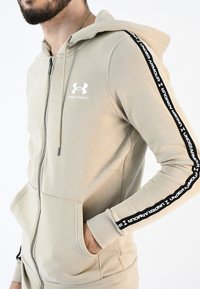 Sudadera con cremallera en color beige, hecha de material suave, que presenta un gran bolsillo frontal y rayas laterales negras con texto y logo en blanco de "Under Armour".