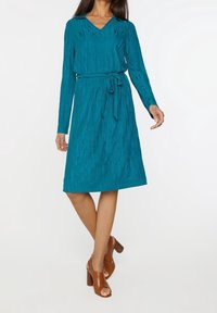 Teal texturerad klänning med V-ringning och långa ärmar, med ett midjeband. Buren med bruna öppetågade klackskor.