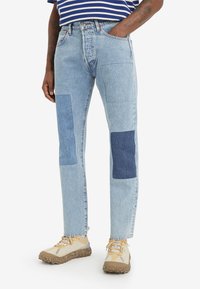 Muž oblečený v světle modrých denimových džínách s tmavomodrými nášivkami na kolenou a béžových teniskách, ruka v kapse.