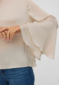 Donna che indossa una blusa beige con maniche a campana a strati e jeans blu, con le mani che si toccano delicatamente all'altezza della vita.