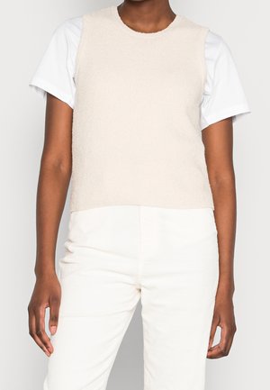 Femme portant un top en maille beige sans manches par-dessus une chemise blanche à manches courtes avec un pantalon blanc sur un fond clair.