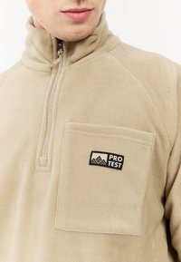 Beige fleecepullover met een hoge kraag, een halve rits aan de voorkant en een zak aan de voorkant met een zwart "PRO TEST" label. Zachte textuur en eenvoudig ontwerp.
