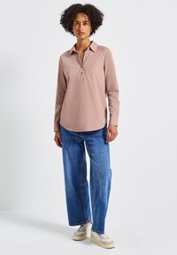 Camicia a maniche lunghe rosa chiaro con colletto bottonato, orlo arrotondato, abbinata a jeans blu larghi e sneakers beige.