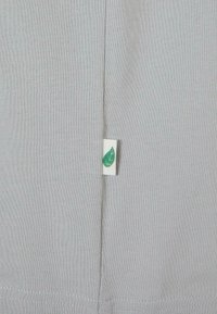 Țesătura gri are o etichetă verde în formă de frunză. Materialul este moale și neted, având o textură subtilă, care pune în evidență un design simplu cu margini clare.
