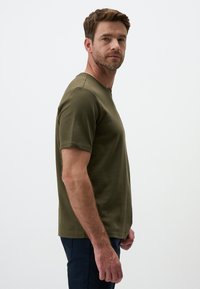 T-shirt verde oliva a maniche corte in cotone, con collo a giro e vestibilità rilassata. Indossato con pantaloni scuri, mostrato in vista laterale.