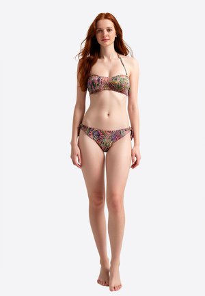 Giovane donna con i capelli lunghi e rossi in piedi, che indossa un bikini multicolore con fantasia e slip con fiocchi laterali su uno sfondo bianco semplice.
