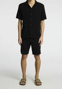 Czarna, krótka koszula na guziki z kołnierzykiem i dopasowane czarne szorty wykonane z teksturowanej tkaniny. Stylizowane z sandałami.