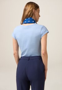 T-shirt in cotone blu chiaro con maniche corte, abbinato a pantaloni navy su misura; un foulard blu con pattern bianchi è visibile all'collo.