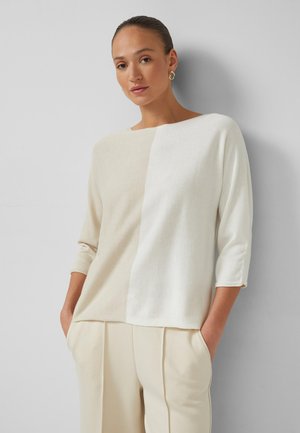 Vrouw met naar achteren gekamd haar, gouden oorringen, een beige-witte kleurvlakken-trui en beige broek met handen in de zakken.