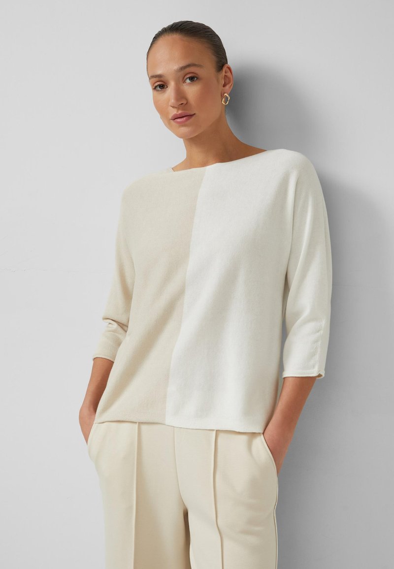 Frau mit zurückgekämmtem Haar, die goldene Creolen, einen beigen und weißen Farbblockpullover sowie beige Hosen trägt und die Hände in die Taschen steckt.