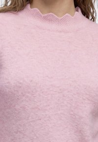Pull en maille rose doux avec un col montant texturé à bord festonné porté par une personne avec des mèches de cheveux châtain clair visibles.