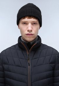 Giacca nera trapuntata con collo alto, dettagli della cerniera arancioni, indossata sopra un maglione grigio a coste e completata da un berretto di maglia nero.