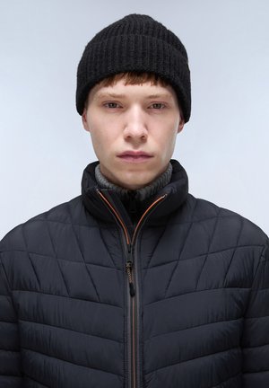 Giacca nera trapuntata con collo alto, dettagli della cerniera arancioni, indossata sopra un maglione grigio a coste e completata da un berretto di maglia nero.