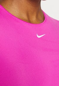 Fuchsiové sportovní tričko s kulatým výstřihem a bílým logem Nike. Obsahuje texturovanou, prodyšnou látku s malými perforacemi.