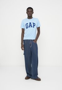 T-shirt bleu clair à col rond avec lettrage "GAP" bleu marine, associé à un pantalon ample rayé bleu marine et des chaussures marron.