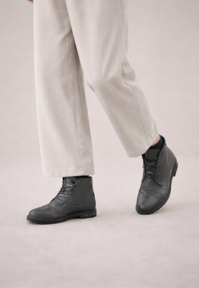 Person wearing light beige pants and black leather lace-up ankle boots standing on a light floor.