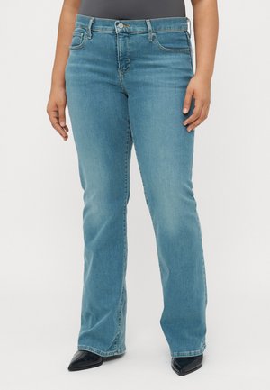 Femme portant un jean bootcut bleu clair avec poches avant et fermeture par boutons, associée à des chaussures noires à bout pointu sur fond blanc.