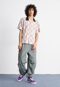 Short-sleeve shirt with a geometric pattern in beige and burgundy, paired with cargo-style green trousers and purple sneakers.