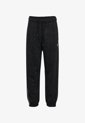 Dziewczyna z warkoczami, ubrana w czarny dres w plamki i białe trampki, stoi i uśmiecha się na jednolitym tle.