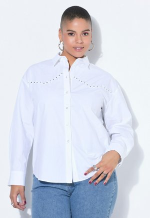 Kvinde med kort hår iført en hvid langærmet skjorte med knapper og nitter og blå jeans, poserer med den ene hånd på hoften.