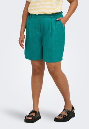 Kvinde iført teal knælange shorts, sorte sandaler med spænde og en gul- og hvidstribet skjorte med hænderne i lommerne.