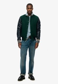 Heren varsityjack met een groene wollen body, zwarte leren mouwen, gestreepte manchetten, knoopsluiting, gedragen over een lichtdenim overhemd en blauwe spijkerbroek.