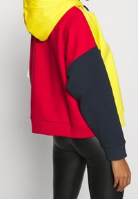 Persona che indossa un hoodie color block con sezioni gialle, rosse e blu navy, abbinato a leggings in pelle nera, in piedi contro uno sfondo neutro.