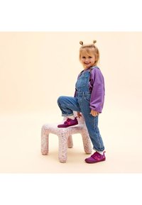 Denim tuinbroeken met verstelbare banden, gecombineerd met een paarse lange mouwen shirt. Kind dat op een gestructureerde, roze en witte gespikkelde kruk staat, met roze schoenen aan.