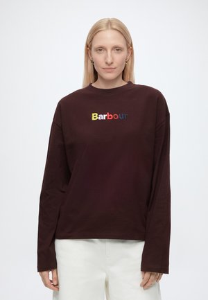 Blonde woman wearing a dark brown long-sleeve shirt with colorful "Barbour" logo, paired with white pants, standing against a plain white background.