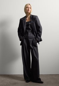 Jeune femme aux cheveux blonds courts, portant un costume sombre ample, une veste et un pantalon large, se tenant devant un mur blanc uni.
