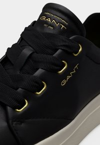Μαύρο δερμάτινο αθλητικό παπούτσι με παχιά μαύρα κορδόνια, χρυσά μεταλλικά τρύπια και λογότυπο "GANT" σε χρυσό στη γλώσσα και στην πλευρά. Φαίνεται λευκή σόλα.