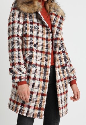 Kvinde iført en dobbeltradet, ternet frakke med sorte, hvide, røde og beige tern, med brun pelskrave og manchetter med knapper.