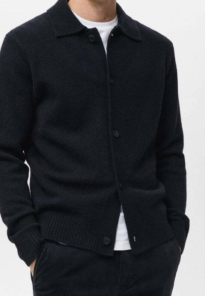 Homme portant un cardigan noir boutonné sur un t-shirt blanc, les mains dans les poches, sur un fond uni.