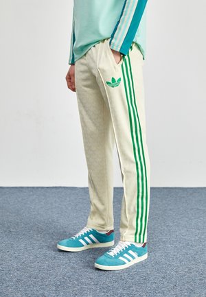 Osoba nosząca kremowe spodnie dresowe Adidas z zielonymi paskami i logo, turkusowe buty sportowe z białymi paskami, ręka w kieszeni, stoi na niebieskiej podłodze.